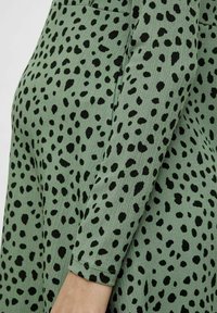 Zelené texturované šaty s nepravidelnými černými puntíky, rukáv a ruka částečně viditelné na spodní části.