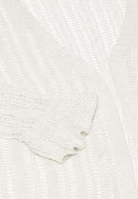 Weißer Strickcardigan mit V-Ausschnitt, geripptem Muster und locker sitzenden Ärmeln mit ausgefransten Kanten.