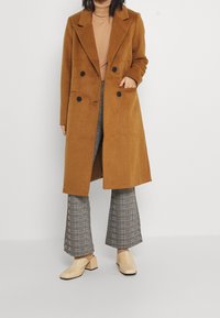 Manteau marron à double boutonnage avec de grands revers, boutons noirs et poches latérales. Associé à un pantalon évasé à carreaux gris et des chaussures à talons blocs beiges.