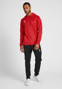 Rote Kapuzenjacke mit schwarzen Akzenten, kombiniert mit schwarzen Jogginghosen mit einem Logo. Weicher, leichter Stoff; ausgestattet mit Kordeln.