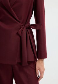 Top avvolgente bordeaux con maniche lunghe, caratterizzato da un dettaglio in vita legato e una texture liscia. Abbinato a pantaloni bordeaux coordinati.