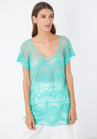 Top turquesa transparente con mangas en forma de scallop, que presenta bordados florales intrincados y un corte relajado. Material suave con una textura ligera.