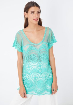 Top turquesa transparente con mangas en forma de scallop, que presenta bordados florales intrincados y un corte relajado. Material suave con una textura ligera.