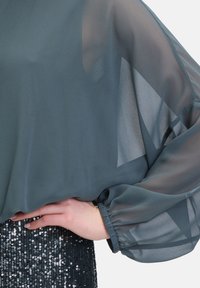 Top in chiffon di un verde scuro con maniche a taglio morbido; presenta polsini elasticizzati. Indossato sopra una gonna decorata con paillettes nelle tonalità di argento e nero.