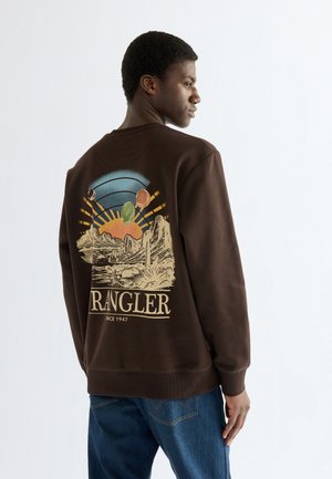 Jeune homme portant un sweat-shirt Wrangler marron avec un paysage désertique coloré et un motif de soleil dans le dos, associé à un jean bleu.