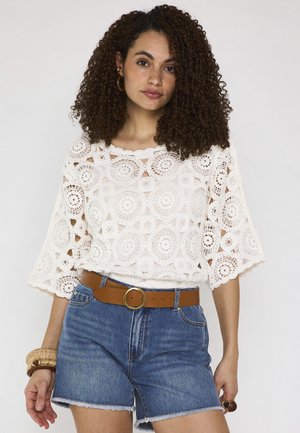 Giovane donna con capelli ricci che indossa un top bianco all'uncinetto, pantaloncini di jeans con cintura marrone e braccialetti al polso sinistro.
