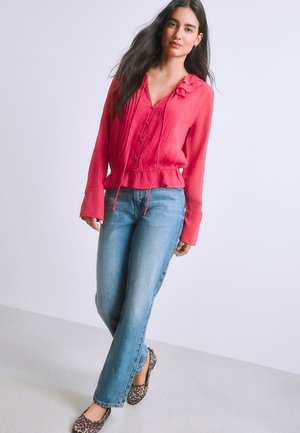 Femme aux longs cheveux foncés portant un chemisier rouge à volants, un jean bleu et des ballerines à imprimé léopard, debout devant un fond uni clair.