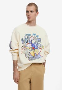 Crème sweatshirt met een kleurrijke afbeelding van een cartoon-eend, tekst met "Sorry voor mijn slechte humeur," en geribbelde manchetten. Tan broek maakt de look compleet.