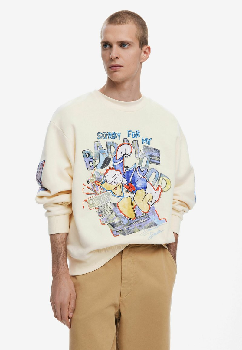 Crème sweatshirt met een kleurrijke afbeelding van een cartoon-eend, tekst met "Sorry voor mijn slechte humeur," en geribbelde manchetten. Tan broek maakt de look compleet.