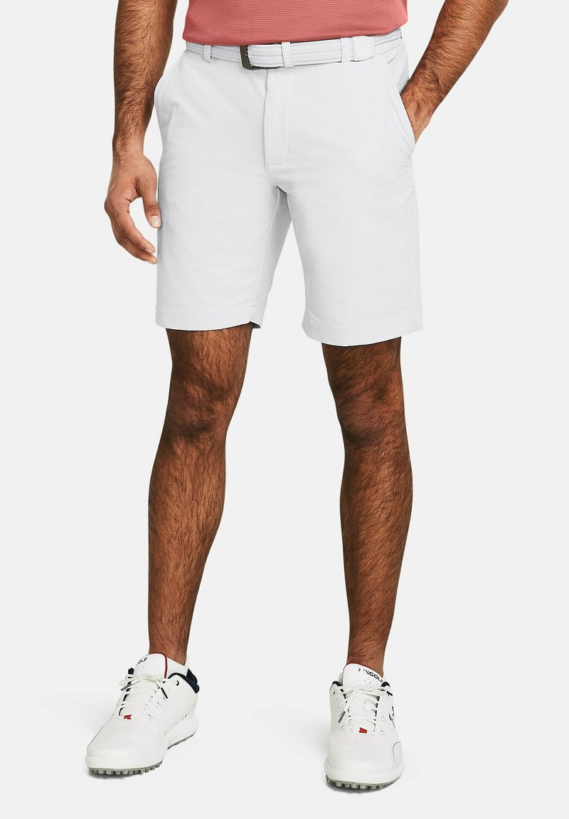 Homme portant un short blanc, une ceinture blanche et des baskets blanches, debout avec une main dans la poche sur un fond uni.