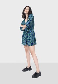 Vestido verde de manga corta con estampado de leopardo azul, cintura fruncida y falda acampanada; combinado con zapatos de plataforma negros brillantes.