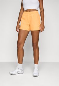 Orange athletic shorts with an elastic waistband, featuring a small logo accent. They are paired with a white crop top and white sneakers.