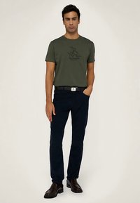 Maglietta verde oliva a maniche corte con motivo grafico nero, abbinata a pantaloni blu scuro e scarpe in pelle nere, che mostrano un outfit casual.