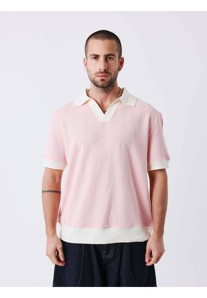 Man met kort haar en baard, die een roze kort m mouw polo met kraag en een donkerbroek draagt, staand tegen een effen lichte achtergrond.