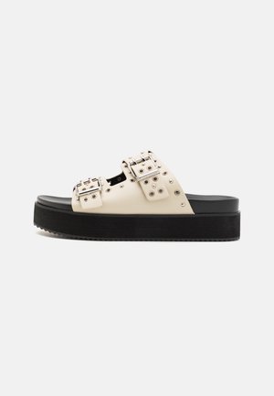 Platformsandaler i off-white kunstlæder med sort sål. Har dobbelte metalspænder og cirkulære nitter som detaljer.