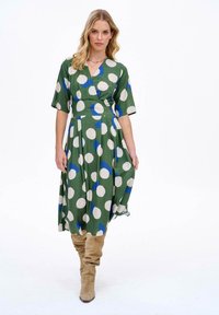 Zelené šaty s velkými bílými a modrými puntíky, zavinovací styl, výstřih do V, krátké rukávy, splývavá sukně, kombinované s béžovými vysokými botami po kolena.