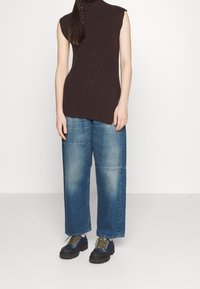 Braunes geripptes ärmelloses Oberteil, kombiniert mit übergroßen hellblauen Jeans mit geradem Schnitt und sichtbaren Nähten. Schwarze Schnürschuhe mit goldenen Akzenten.
