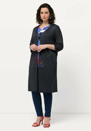 Zwarte, lange cardigan met 3/4 mouwen, open voorkant en zachte textuur. In combinatie met een blauw patronen top en donkerblauwe jeans, met rode hakken.