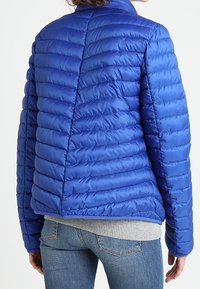 Polstret blå jakke med en let, blank finish, der har vandrette quiltede linjer, krave og elastiske manchetter, båret over en grå sweater.