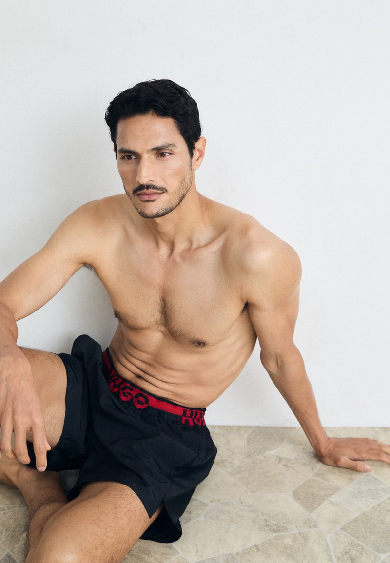Fekete rövidnadrág vörös derékkal, rajta "HUGO" felirattal, egy mezítelen férfi modellen, aki egy textúrázott világos színű padlón ül.