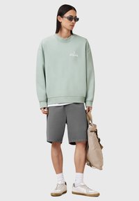 Sudadera verde clara con logo, pantalones cortos grises, calcetines blancos y zapatillas beige. El modelo sostiene un bolso beige, que presenta un ajuste relajado y un diseño sencillo.