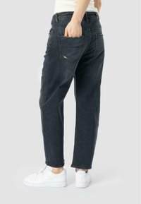 Jean en denim foncé avec une coupe décontractée, présentant des détails déchirés, des poches arrière et des bords usés, associés à des baskets blanches.