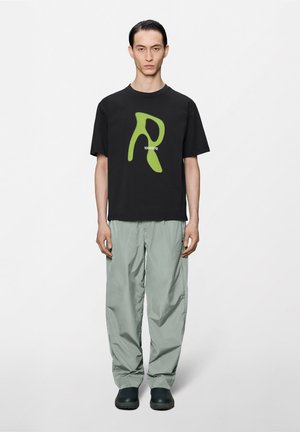 Muž v čiernom tričku s veľkým zeleným písmenom "R" a číslami, v svetlosivých nohaviciach a čiernych topánkach, stojaci pred jednoduchým svetlým pozadím.