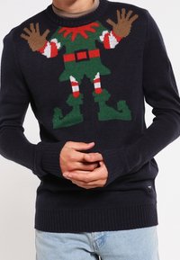 Pull en tricot bleu marine arborant un motif festif d'elfe en vert, rouge et blanc avec des mains détaillées, une ceinture et des manches rayées.