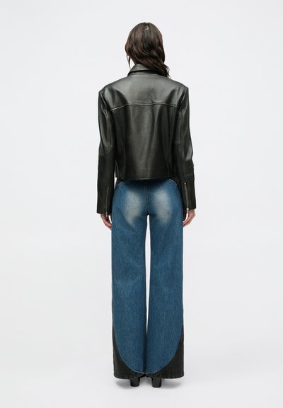 Giacca corta in pelle nera con texture liscia e chiusura frontale con zip, abbinata a jeans di jeans blu a gamba larga con dettagli scoloriti.