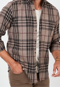 Hnědá kostkovaná flanelová košile s černými a burgundskými pruhy, s knoflíkovým zapínáním a ohrnutými rukávy. Nositá přes světle zbarvenou košili.