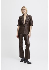Blazer en cuir marron à manches courtes et décolleté en V profond, associé à un pantalon en cuir marron assorti. Des bottes à talons noires complètent la tenue.