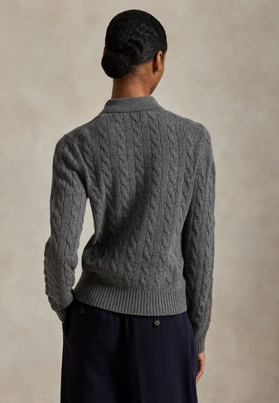 Nainen, jolla on tummat hiukset nutturalla, pukeutuneena harmaaseen palmikkoneulepaitaan ja tummiin housuihin, seisoo selin neutraalia taustaa vasten.