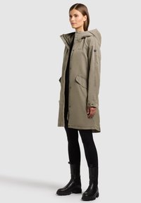 Femme portant une parka à capuche kaki, des leggings noirs et des bottines noires, debout de profil devant un fond clair uni.