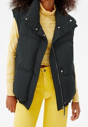 Woman wearing a black padded vest over a yellow patterned turtleneck and high-waisted yellow pants, standing with hands relaxed.