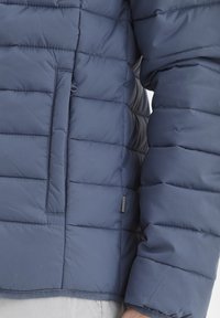 Tumši zila pūkaina jaka ar izšūtu rakstu, ar rāvējslēdzēja kabatu priekšā un mazu birku sānu apakšmalā, mīksta tekstūra.