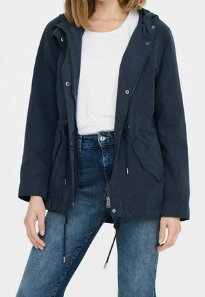 Femme portant une veste à capuche bleu marine par-dessus une chemise blanche et un jean bleu, debout les mains détendues le long du corps sur un fond blanc.