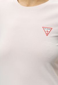 T-shirt blanc avec un petit logo triangulaire Guess bordé de rouge sur la partie supérieure gauche de la poitrine.