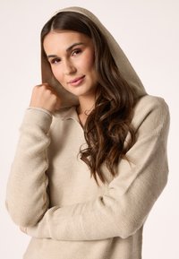 Felpa con cappuccio lavorata a maglia beige, con un design testurizzato, maniche lunghe e cappuccio regolabile. Il modello ha capelli castani ondulati e un'espressione neutra.