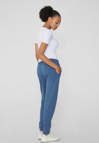 Frau mit lockigem Haar, die ein weißes, eng anliegendes T-Shirt und blaue Jogginghosen trägt. Sie steht mit Händen in den Taschen und schaut nach rechts unten.