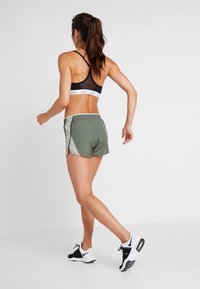 Pantalones cortos deportivos verdes con aberturas laterales, combinados con un sujetador deportivo negro que presenta una banda con logo blanco. La modelo lleva zapatillas negras y blancas.