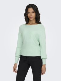 Frau mit dunklem, welligem Haar, die einen hellgrünen, gerippten Pullover und schwarze Hosen trägt, steht vor einem schlichten hellgrauen Hintergrund.