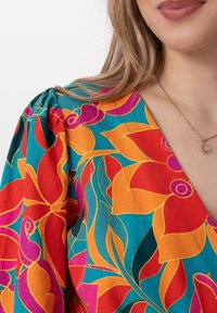 Blouse florale colorée avec des motifs rouges, oranges et violets audacieux sur un fond teal. Comprend un décolleté en V et des manches bouffantes.