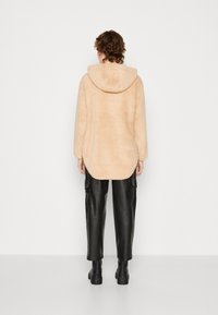 Giacca in pile beige, morbida, con cappuccio, maniche lunghe e orlo curvo. Abbinata a pantaloni cargo neri a vita alta e stivali neri chunky.