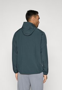 Tumši zaļa kapucēta jaka ar gludu tekstūru, elastīgiem aprocēm un vaļēju piegriezumu, attēlota no aizmugures. Viegls dizains, piemērots lietošanai ārpus telpām.