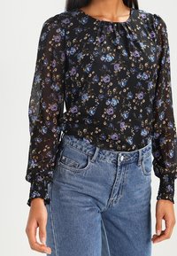 Donna che indossa una blusa floreale nera trasparente con fiori viola e blu e jeans blu a vita alta, in piedi con le braccia rilassate.