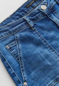 Denim shorts in een middentint blauwe kleur met een verweerd uiterlijk, voorzien van een knoopsluiting, metalen accenten en schuine voorkeurs.