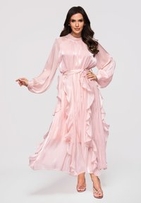 Femme en robe longue fluide et brillante de couleur rose pâle à manches longues avec des volants, taille cintrée, posant avec une main sur la hanche et l'autre tendue.