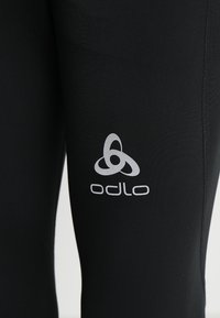 Schwarze Sportleggings aus dehnbarem Stoff, mit einem silbernen Odlo-Logo auf dem Oberschenkel. Glatte Textur und enganliegendes Design.