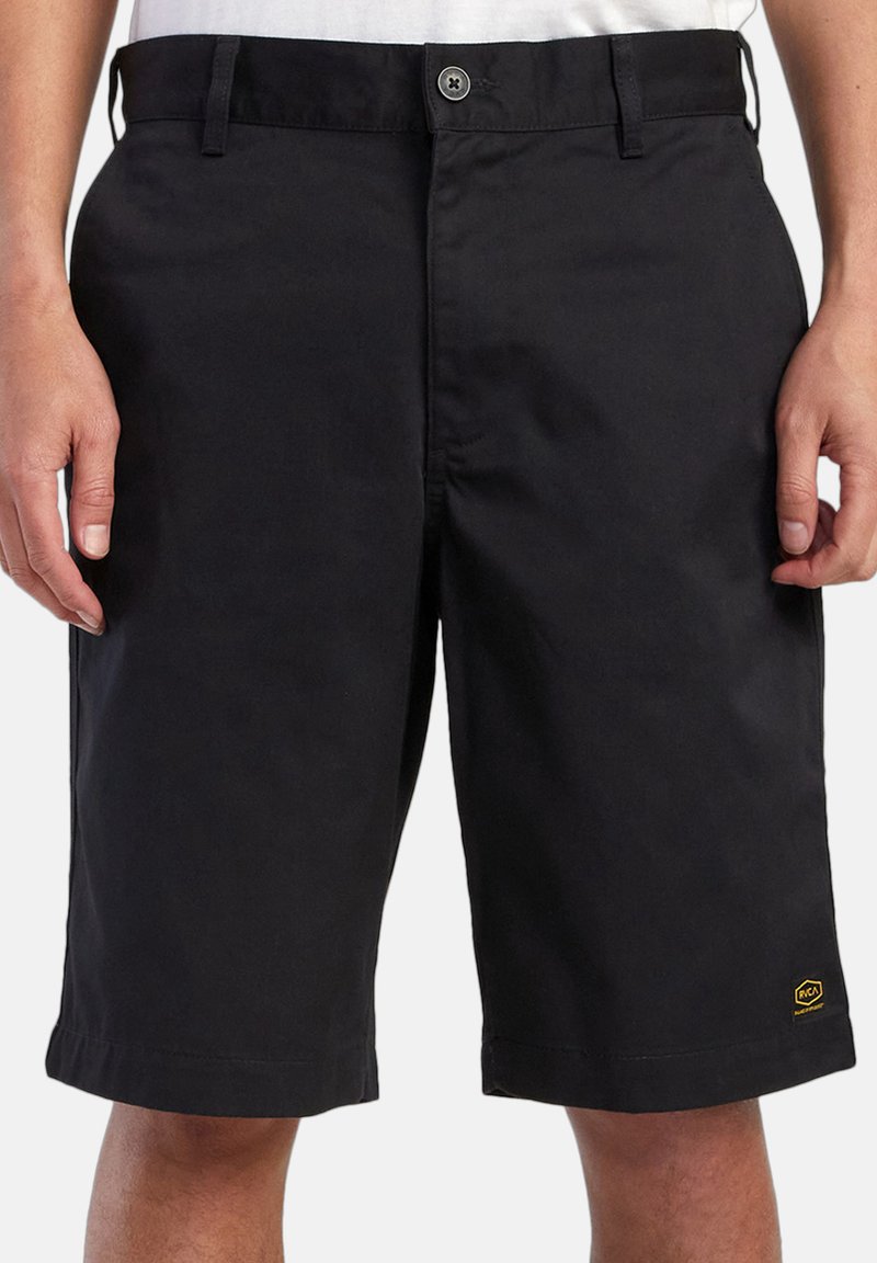 Pantalones cortos negros hasta la rodilla con cierre de botón y cremallera, usados por una persona con las manos a los lados, pequeño logo amarillo en el dobladillo izquierdo.