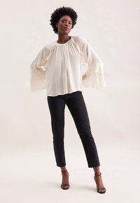 Kremowa bluzka z falbanianymi, rozkloszowanymi rękawami; gładki materiał; w parze z ciemnymi, dopasowanymi jeansami; modelka stoi pewnie na neutralnym tle.